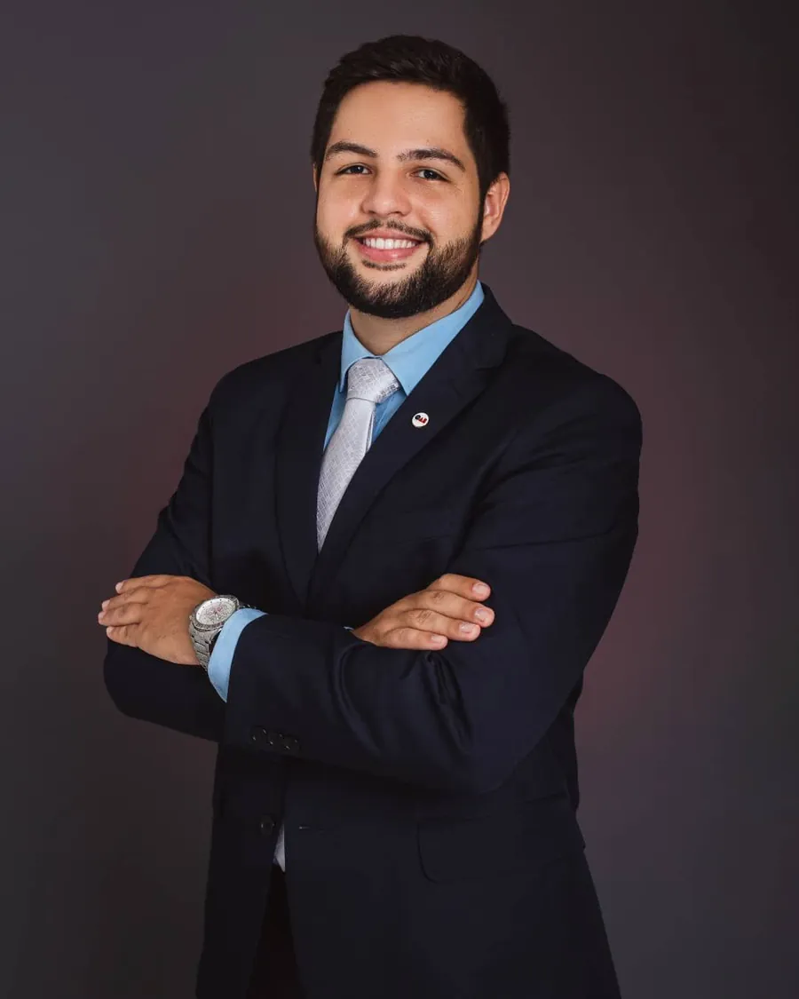 Matheus Aragão de terno azul-marinho e braços cruzados, sorrindo, em fundo escuro